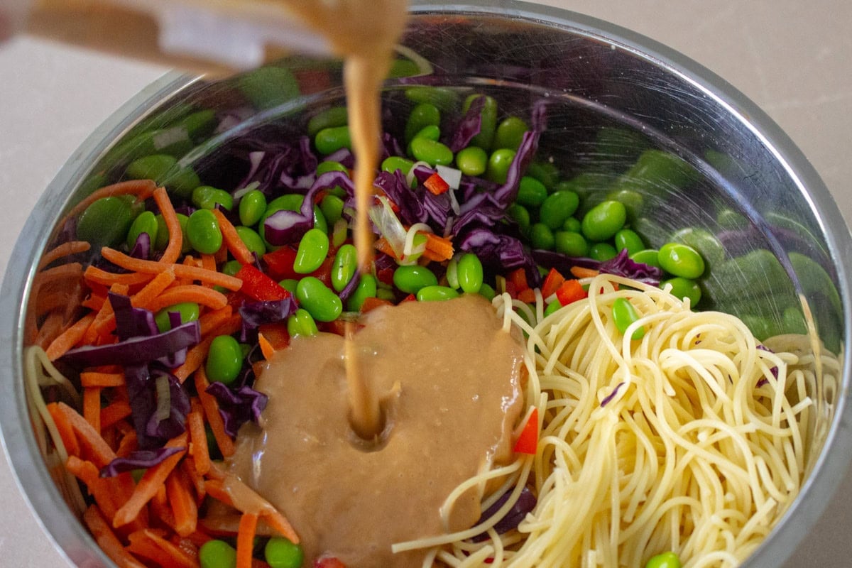 Peanut noodles ingredients in bowl with peanut sauce pouring on top.