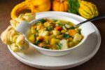 bowl of root vegetable and barley soup with gourds in background. Bowl on plate with bread