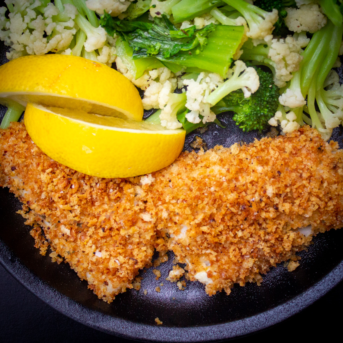 panko covered white fish with veggies on black plate.