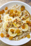 bowl of oatmeal topped with bananas raisins maple syrup nuts p