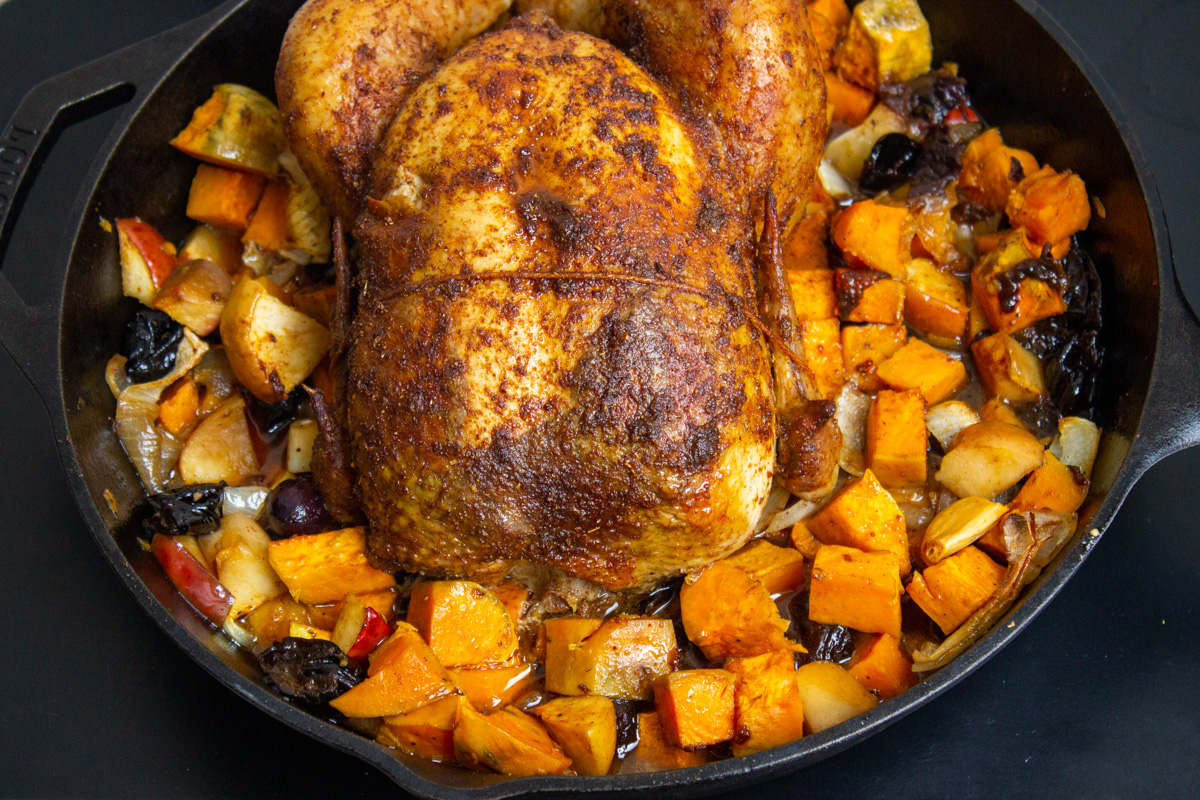 roasted whole chicken nestled on top of cooked veggies and fruit in skillet