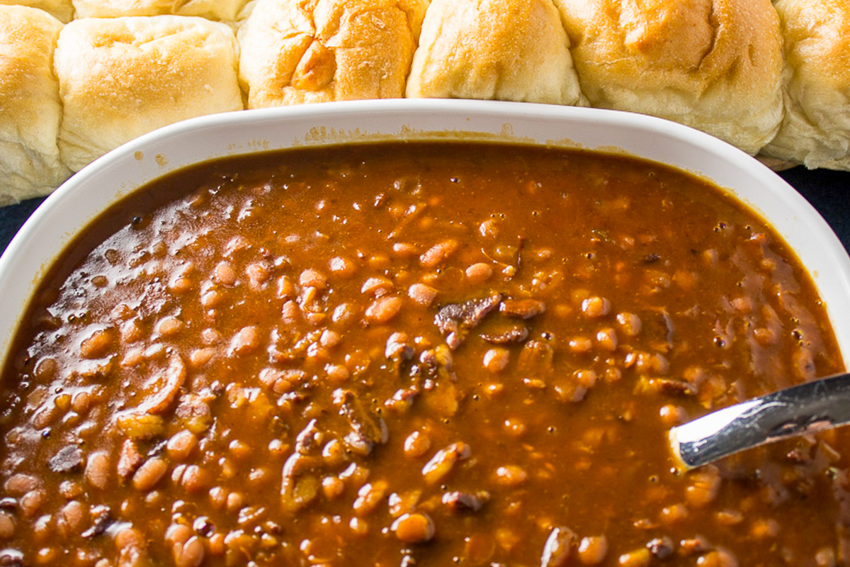 baked beans in serving dish with rolls behind them.