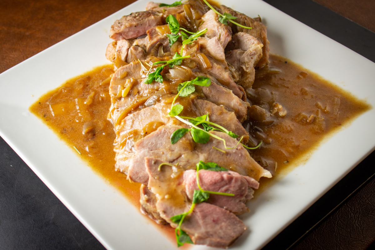 sliced pork roast with gravy on white plate with pea shoot garnish.