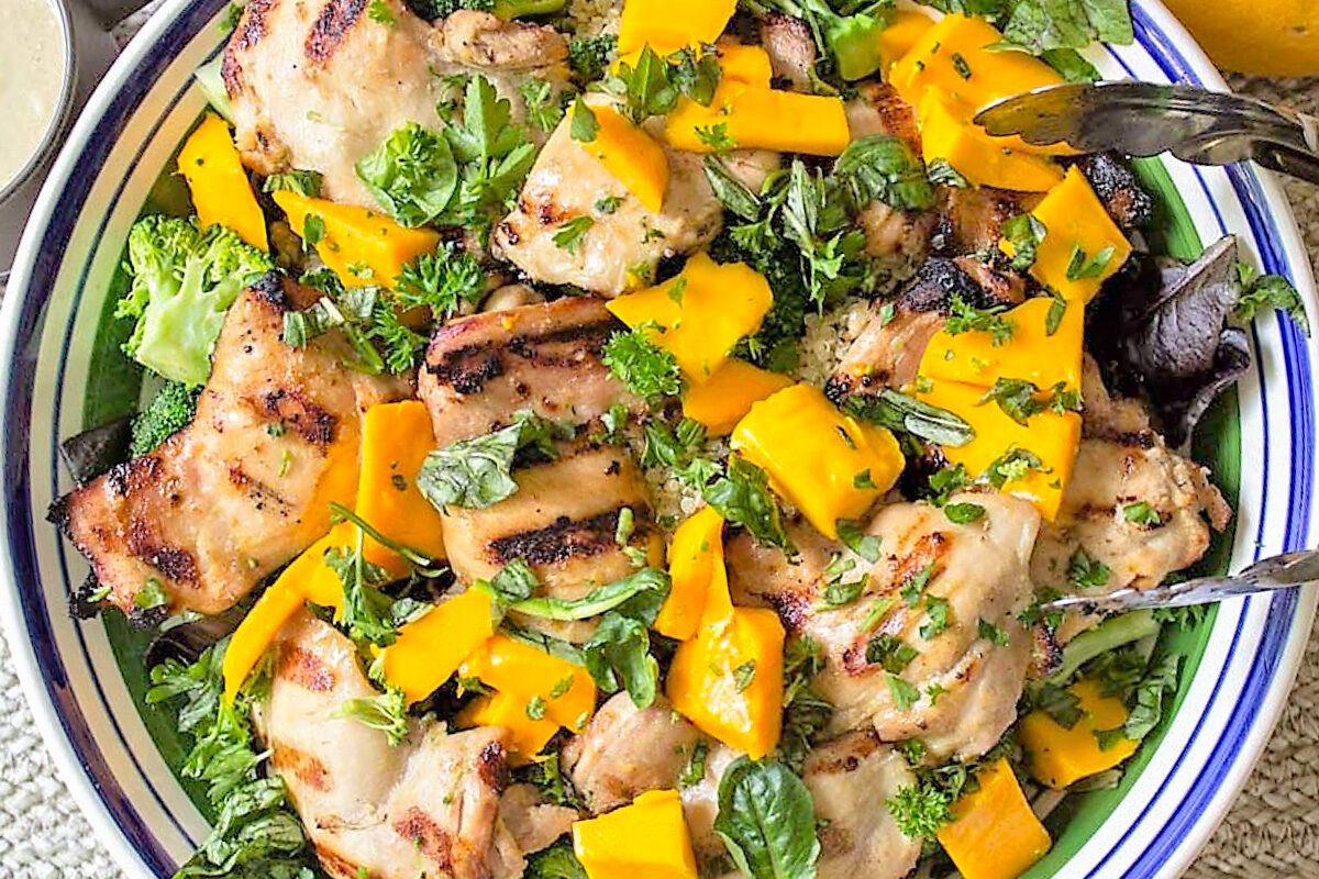 grilled thai chicken thighs cut up into a salad with veggies, greens and mango in bowl.