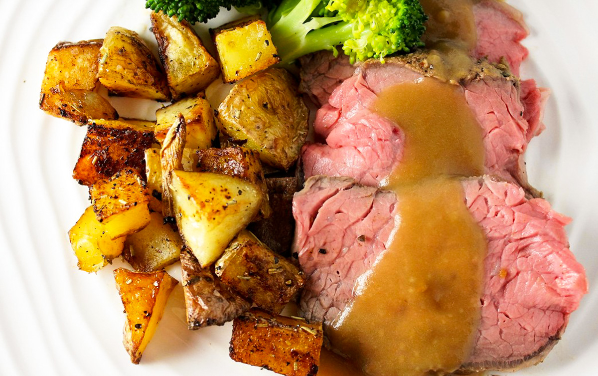 sliced prime rib roast with roast potatoes, gravy and broccoli on plate.