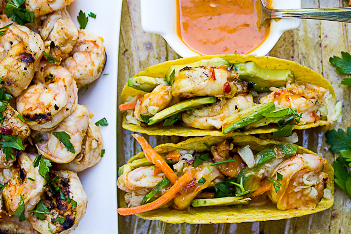 two hard shell coconut shrimp tacos with bowl of sauce and plate of grilled shrimp.