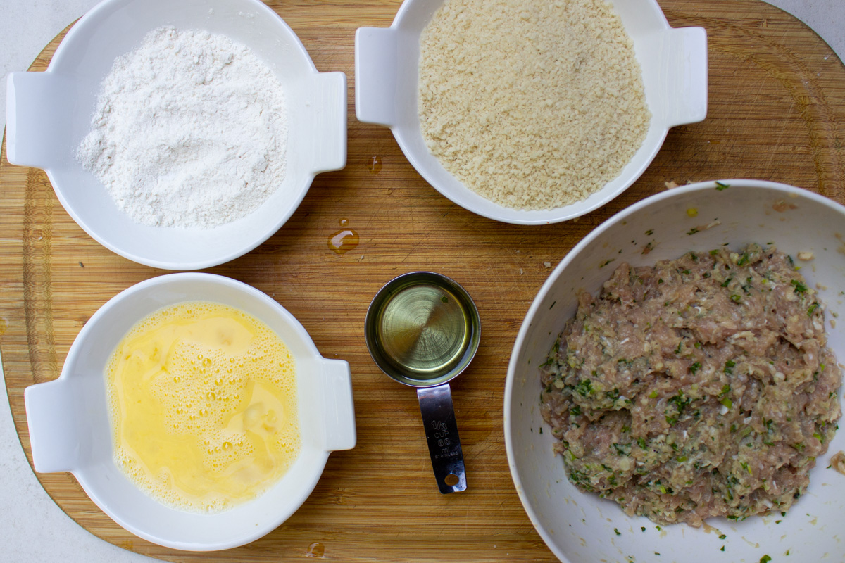 bowls of flour, beaten egg and panko beside oil and ground chicken mixture.