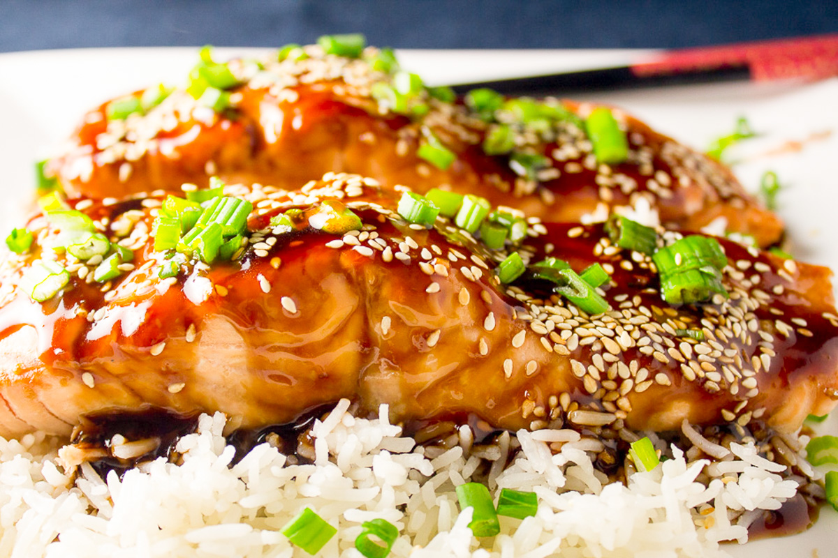 two teriyaki glazed salmon fillets over white rice topped with green onion and sesame seeds on plate..