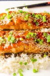 two teriyaki glazed salmon fillets over white rice topped with green onion and sesame seeds on plate..