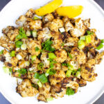 grilled cauliflower garnished with green onions on plate with lemon wedges.