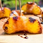 prosciutto-wrapped peaches with toothpicks on wood serving tray with balsamic drizzle.