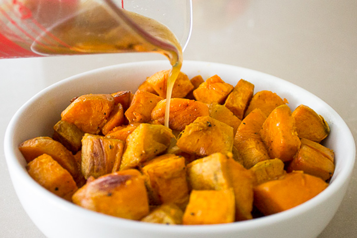 maple syrup mixture pouring onto roasted sweet potatoes in white bowl.