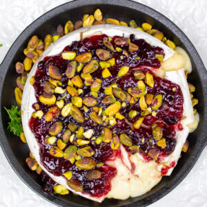 baked brie cheese topped with preserves and pistachios on black plate.