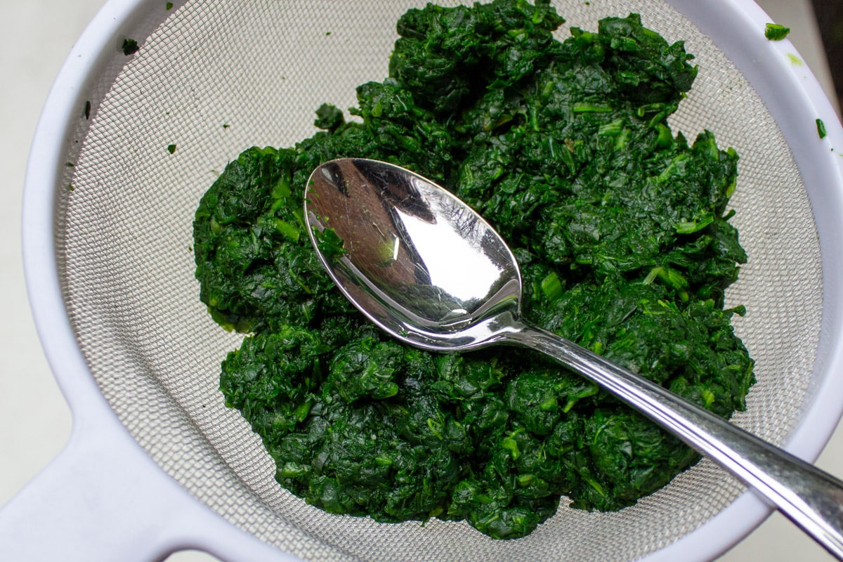 cooked spinach in sieve pressed with spoon to extract water.