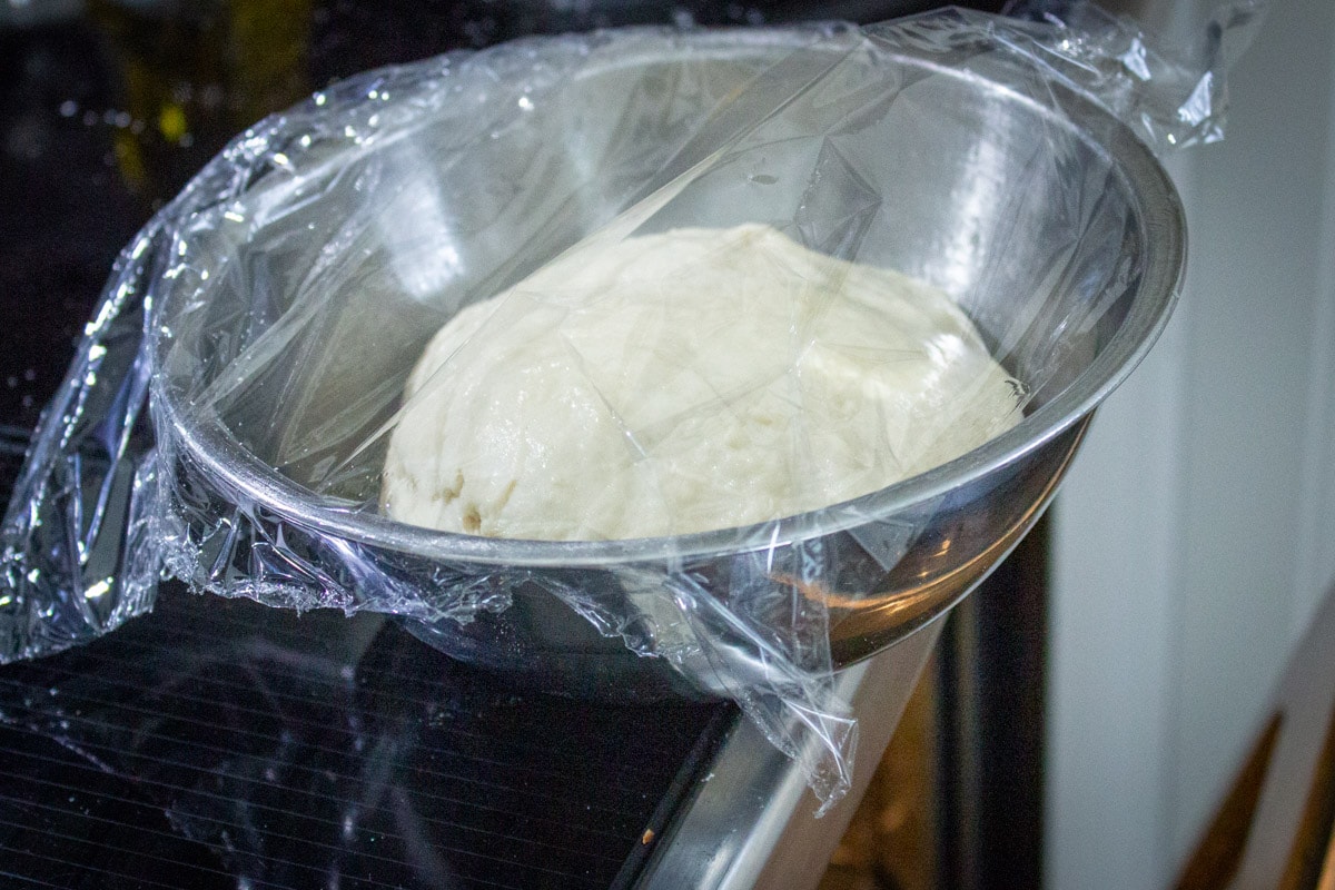 ball of dough in bowl loosely covered in plastic on stove with oven door open.