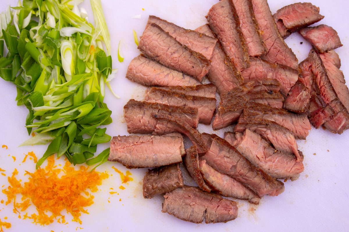 sliced sous vide steak, sliced green onions, shredded carrots on cutting board.
