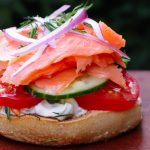 Half bagel piled with cucumber, tomato, lox, red onion and dill on a cutting board.