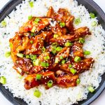 Bourbon chicken garnished with green onion and sesame seeds over white rice on plate.