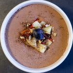 A bowl of chocolate mousse topped with diced apple, toasted nuts, chocolate shavings and blueberries.