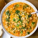 Vegetable and barley soup in a bowl with bread beside it.