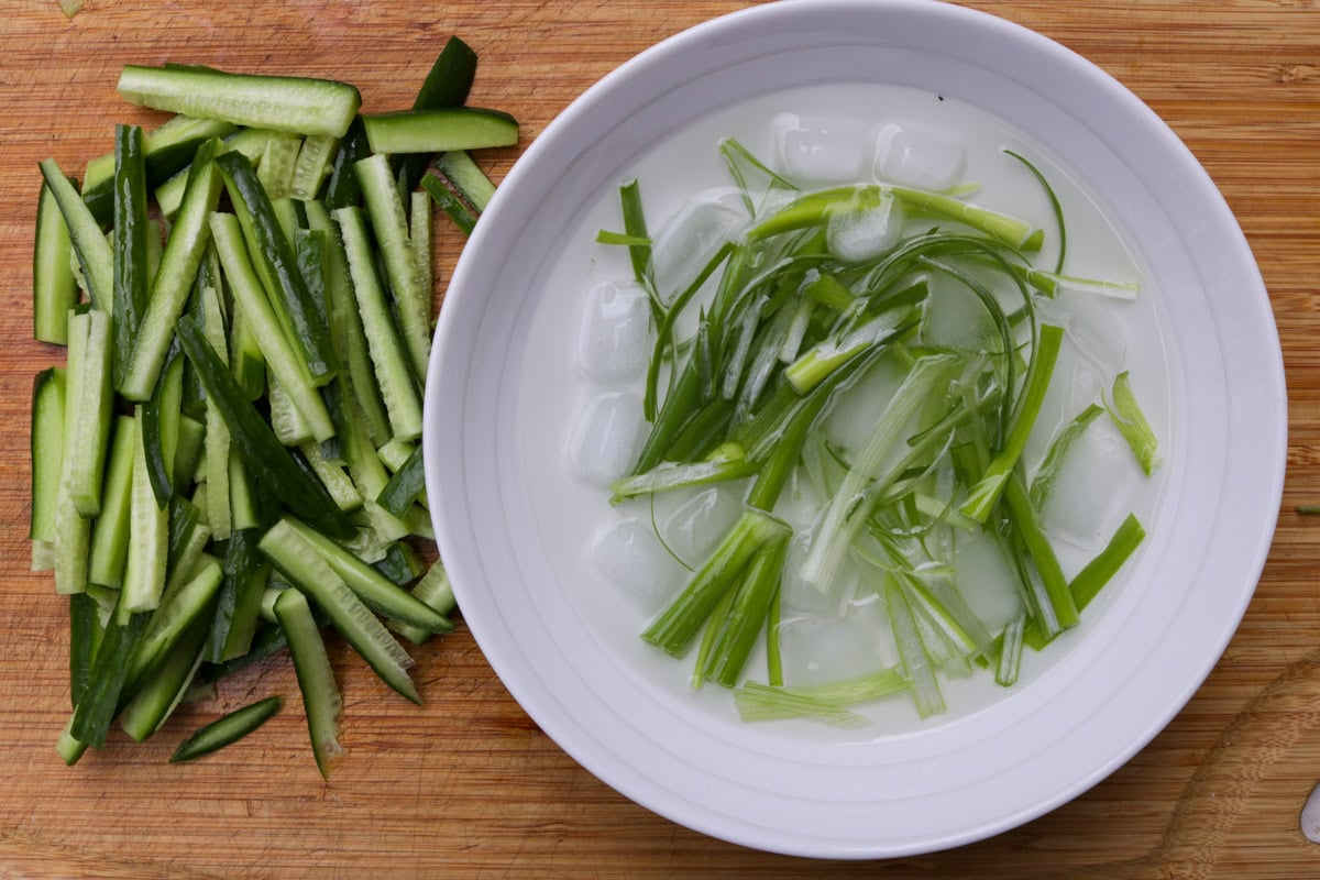 Julienned cucumbers and a bowl of slivered green onions in ice water on a cutting board.