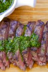 Sliced flat iron steak on cutting board drizzled with chimichurri sauce and bowl of sauce on side.