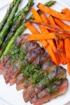 Sliced flat iron steak on cutting board drizzled with chimichurri sauce with sides of sweet potato fries and grilled asparagus.