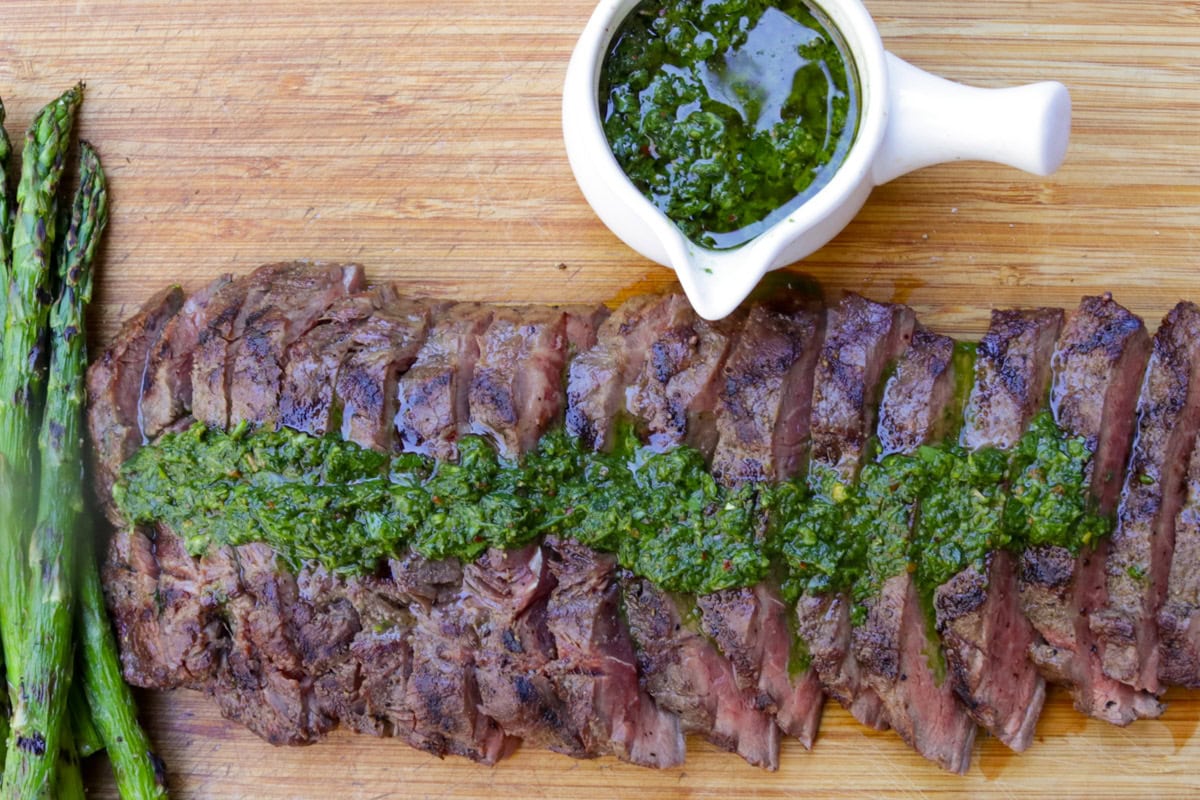 Sliced flat iron steak on cutting board drizzled with chimichurri sauce and bowl of sauce on side.