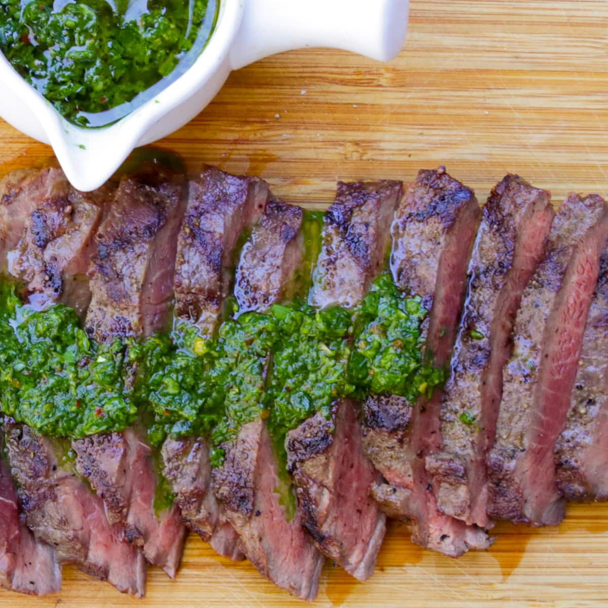 Sliced flat iron steak on cutting board drizzled with chimichurri sauce and bowl of sauce on side.