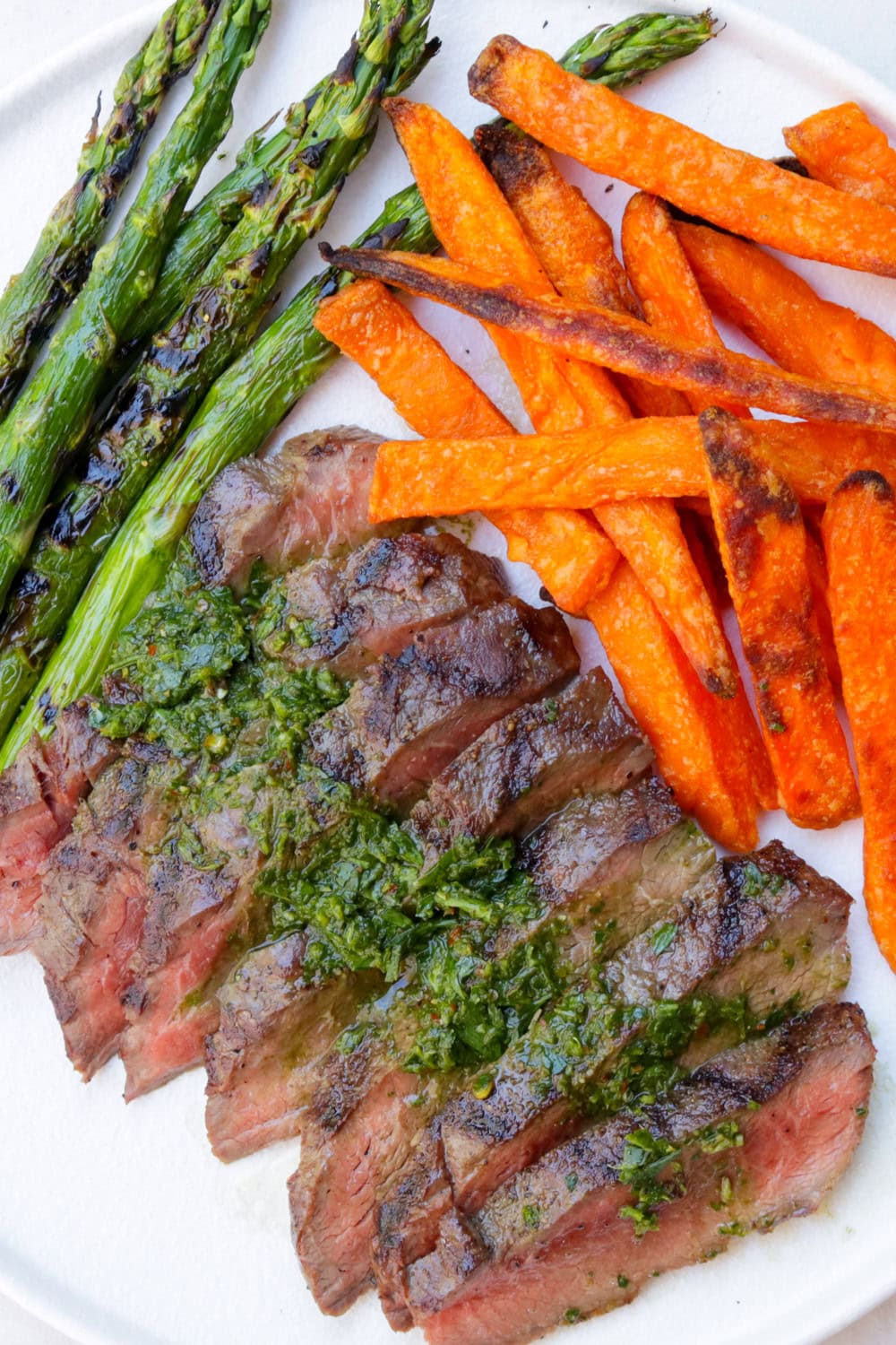 Sliced flat iron steak on cutting board drizzled with chimichurri sauce with sides of sweet potato fries and grilled asparagus.