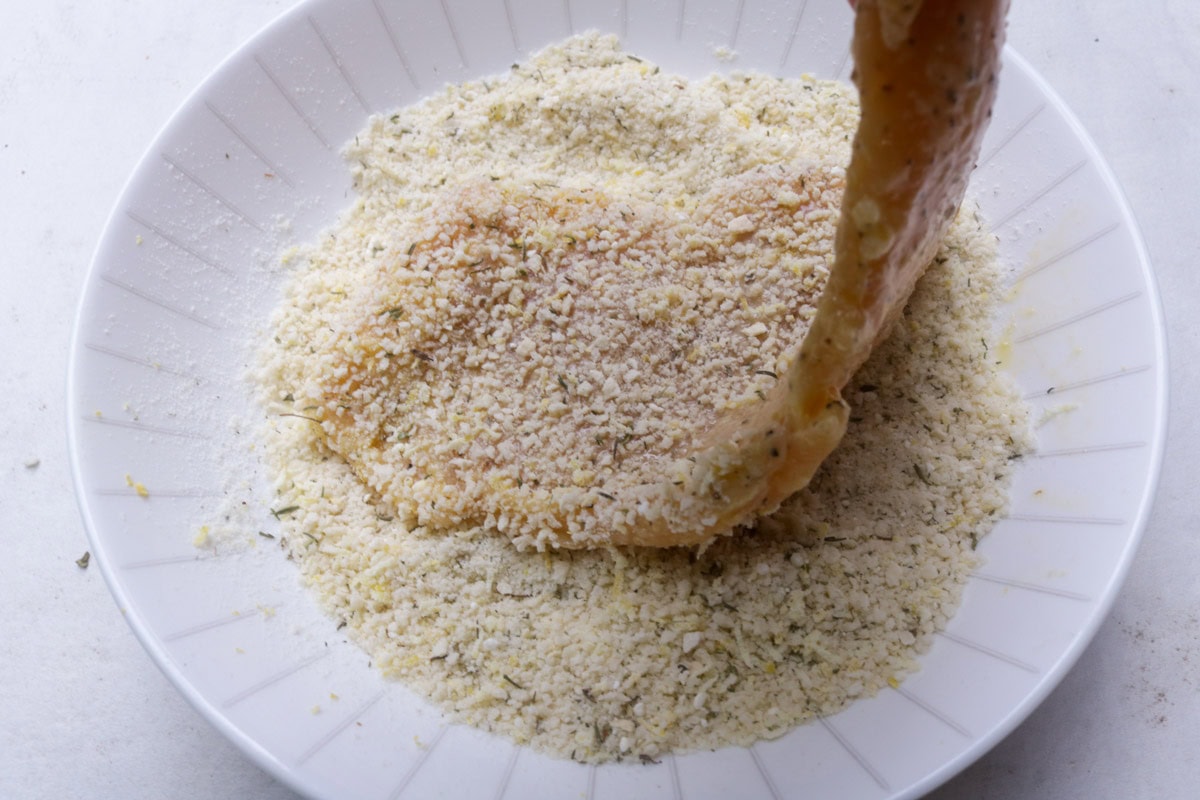 Chicken cutlet coated in flour and eggwash being dipped in panko crumbs in bowl.