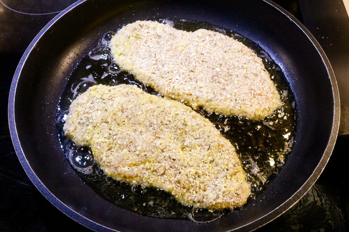 Two breaded chicken cutlets shallow frying in oil in skillet.