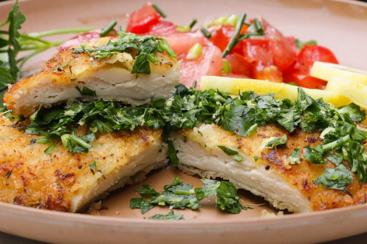 Cut open cooked chicken cutlet with gremolata with lemon slides and tomato salad on plate.