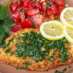 Breaded Italian chicken cutlet with gremolata, tomato salad and lemon slices on plate.