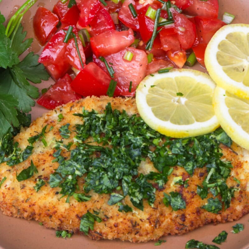 Breaded Italian chicken cutlet with gremolata, tomato salad and lemon slices on plate.