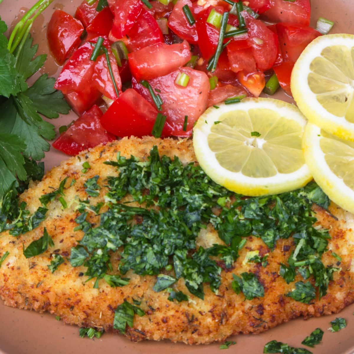 Breaded Italian chicken cutlet with gremolata, tomato salad and lemon slices on plate.