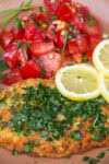Breaded Italian chicken cutlet with gremolata, tomato salad and lemon slices on plate.