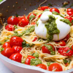 Pasta with tomato mixture in pan with a burrata on top, all drizzled with pesto.