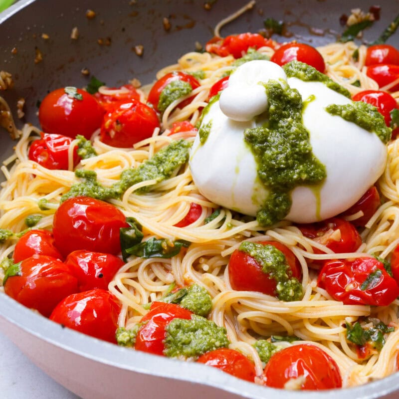 Pasta with tomato mixture in pan with a burrata on top, all drizzled with pesto.