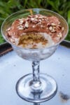 Tiramisu topped with nuts with a scoop missing from it in a glass dessert cup sitting on a plate.