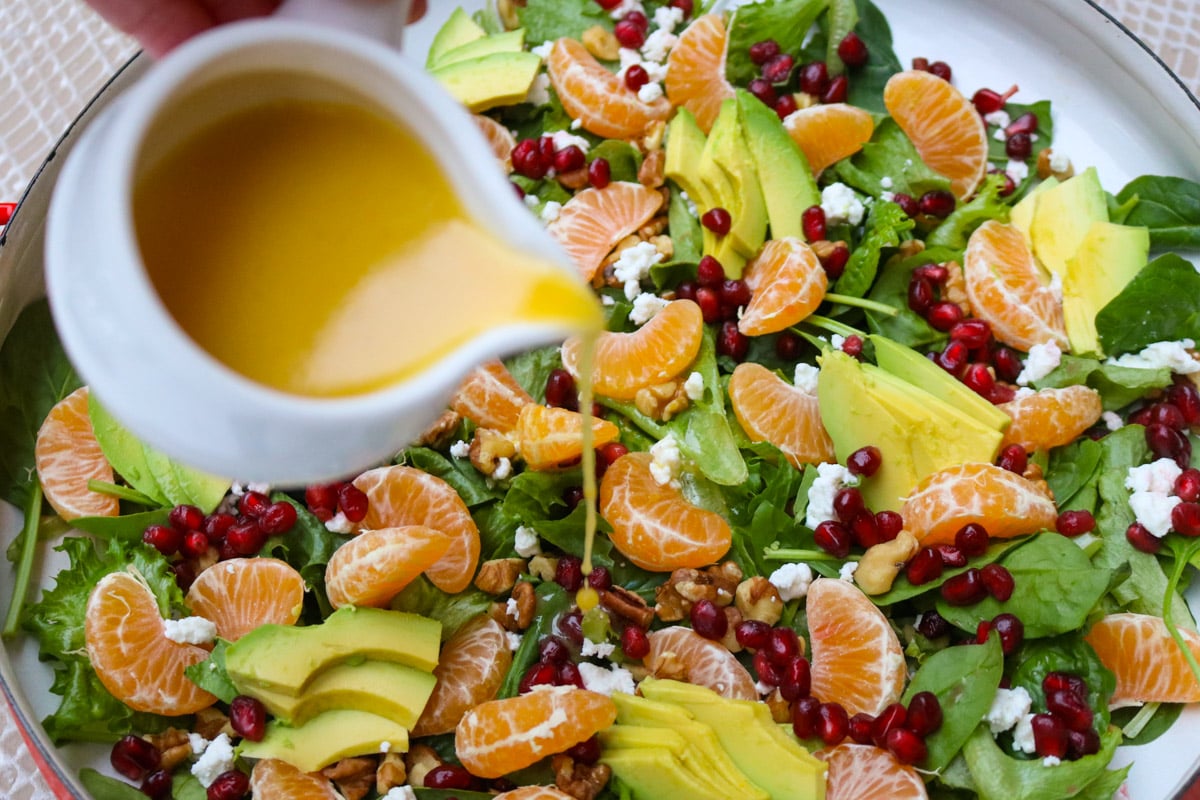 Vinaigrette dressing pouring on a mandarin orange salad with nuts, goat cheese, avocado and pomegranate seeds.