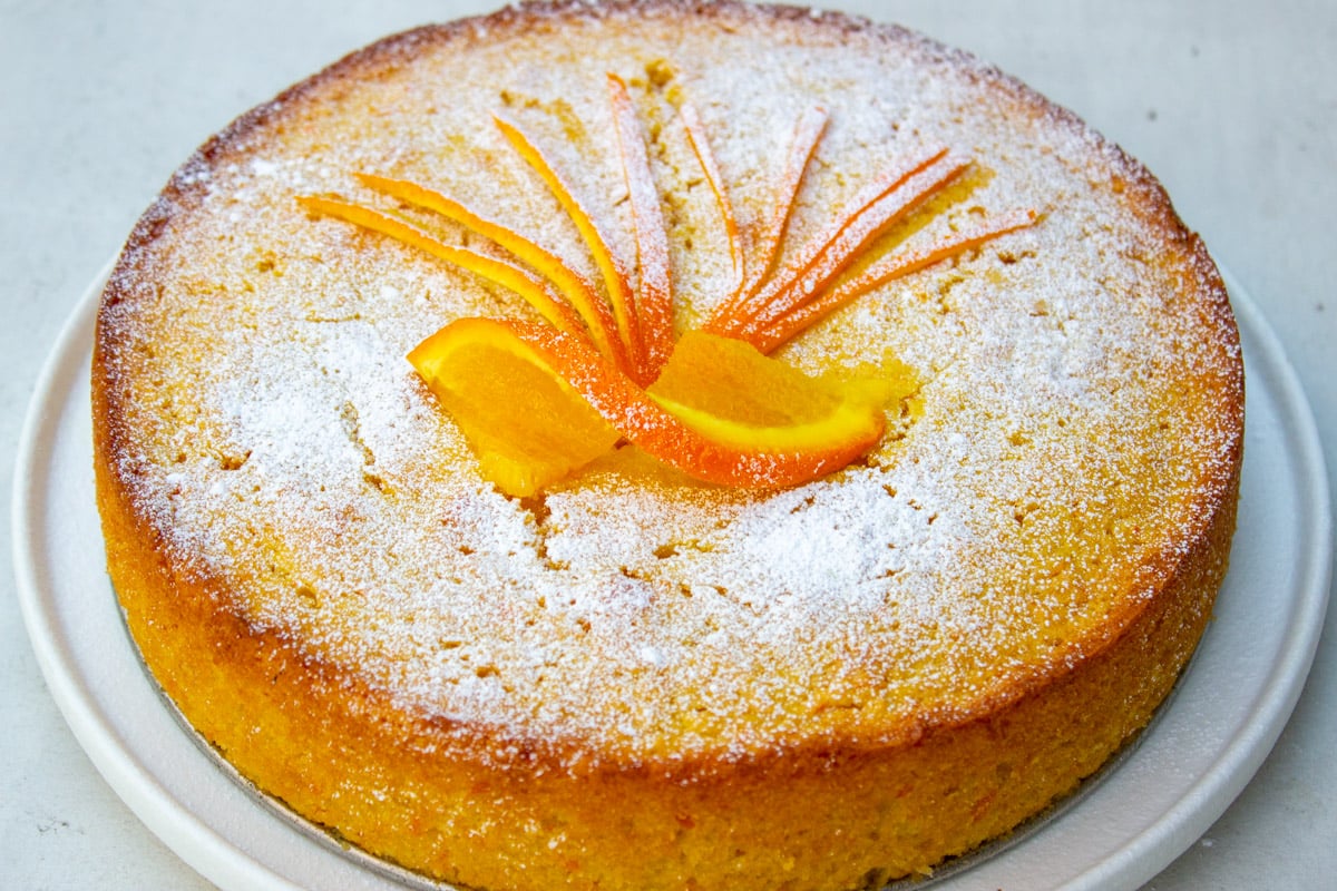 Orange cake dusted with icing sugar and decorated with orange slices and rind.