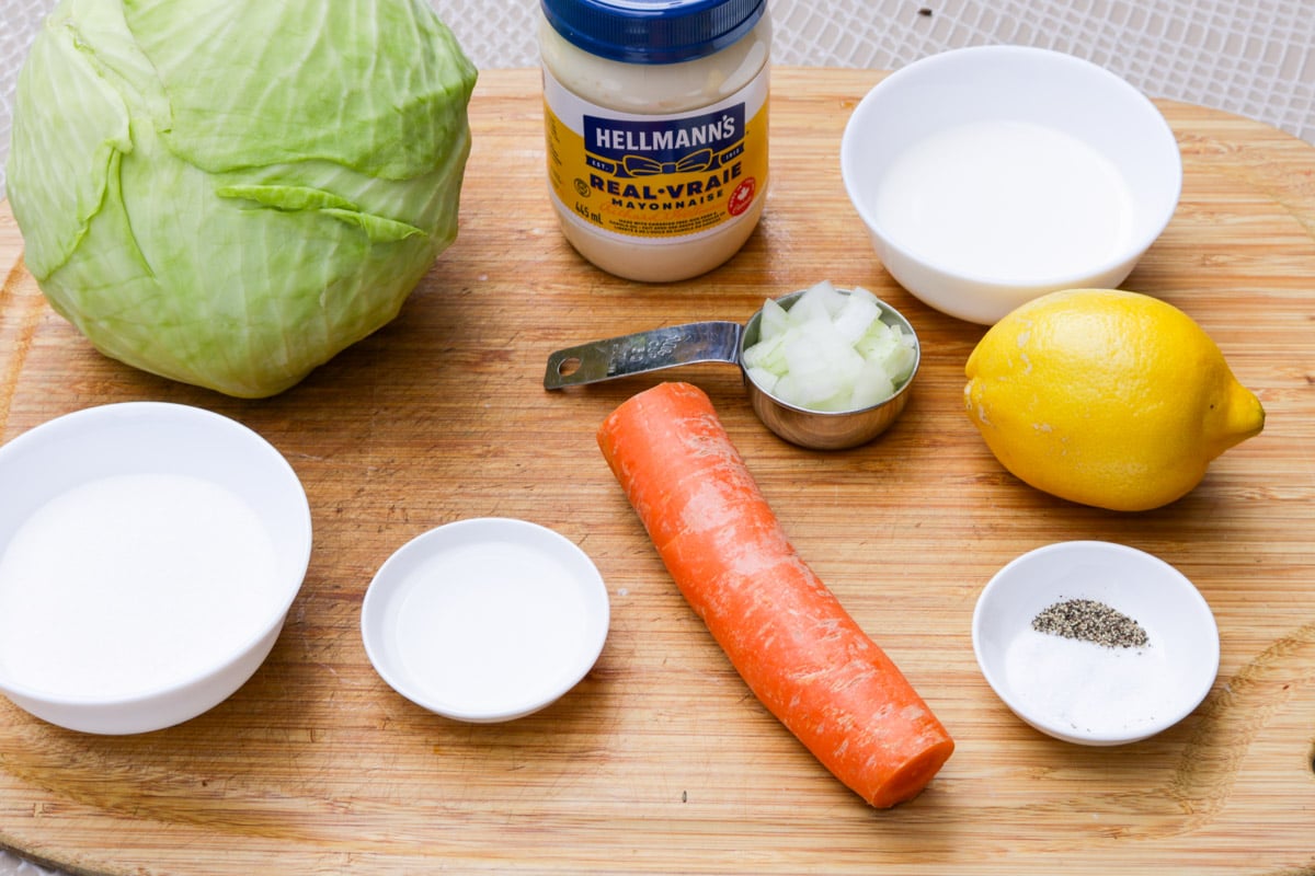 Cabbage, mayonnaise, sugar, milk, lemon, white vinegar, onion, carrot, salt, pepper on cutting board.