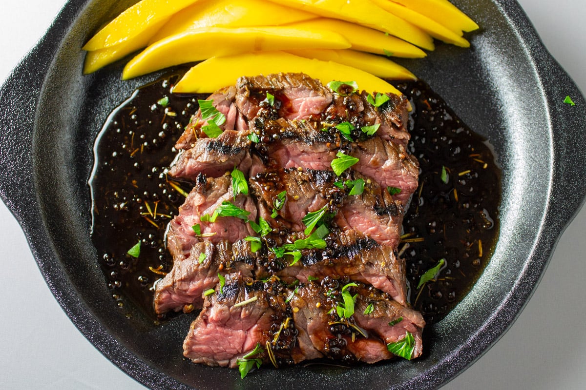 Sliced flank steak with glaze underneath and sliced mango at the top of the plate. 