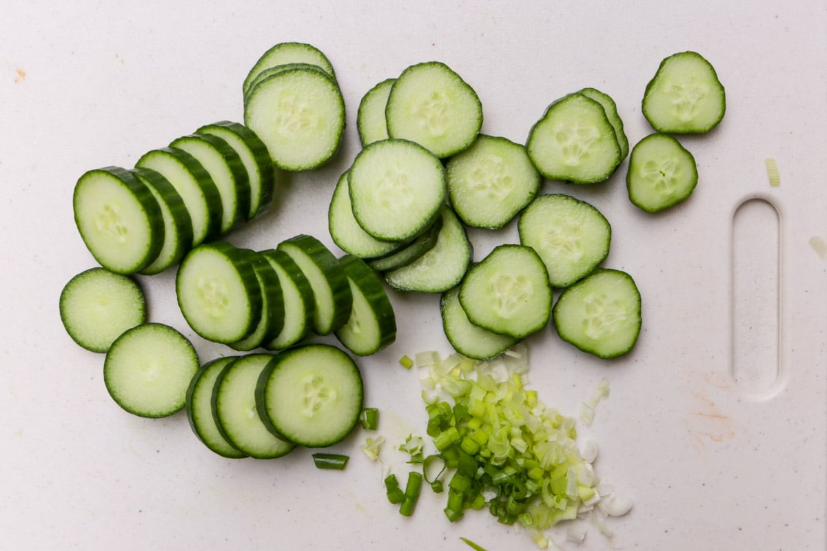 Sliced cucumbers and chopped green onion on white cutting board.
