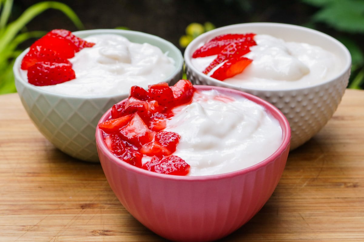 Three individual cups of vanilla pudding with a fresh macerated strawberries on one side of the front bowl.,