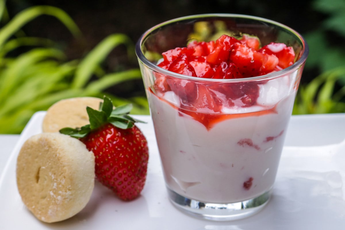 One glass cup of vanilla pudding mixed with strawberries with fresh macerated strawberries on top - with cookies and a strawberry beside it.