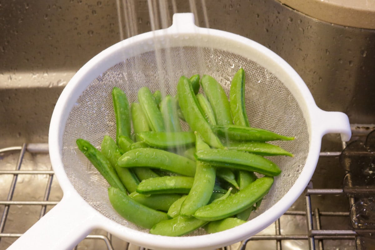 Snap peas in a mesh sieve under spray of water over sink.