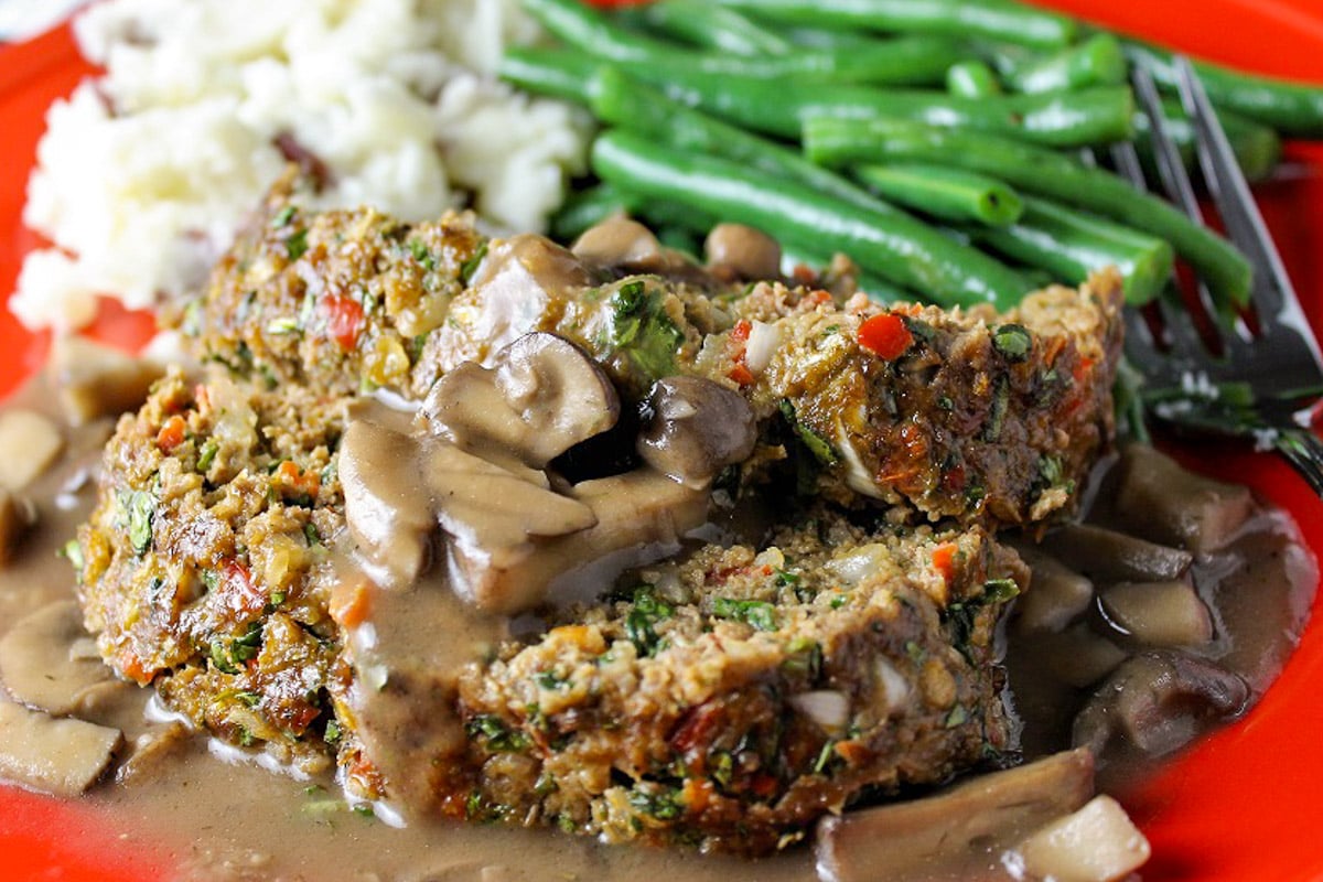 Two slices meatloaf with mushroom gravy, mashed potatoes and green beans on red plate.
