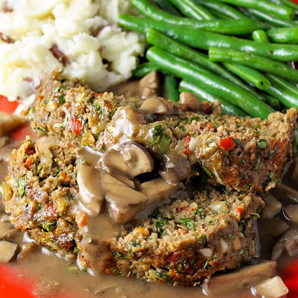 Two slices meatloaf with mushroom gravy, mashed potatoes and green beans on red plate.